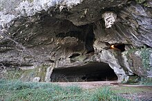 Vista da entrada de uma caverna, abrindo-se como uma boca em um penhasco de calcário cinza claro.