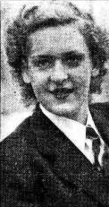 אישה לבנה צעירה עם שיער גלי בלונדיני קצר, לבושה במעיל, חולצה ועניבה.