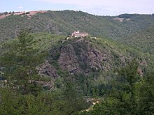 Fotografía en color de una aldea con un campanario en la cima de una cresta rocosa rodeada de empinadas laderas cubiertas de bosque.