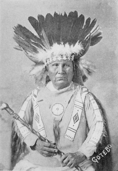 Formal portrait of an elder Kiowa man in traditional dress, including a war bonnet