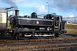 A pannier tank locomotive is pushing a cream and brown passenger carriage. The locomotive is mainly painted black, but there is red and white lining on the side of the pannier tank, the cab, and the bunker. On the side of the pannier tank is a crest consisting of a red and white spoked wheel. Across the wheel is a black and white label with the words "BRITISH RAILWAYS". A yellow lion stands on the label, above the wheel. On the side of the cab is a black number plate with the edge and number in brass. The front buffer beam is red.