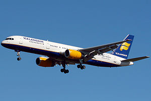 A mostly white Boeing 757 with blue and yellow trim preparing for landing against a blue sky. Landing gear and flaps are fully extended in final approach configuration.