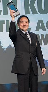 Ken Kutaragi pictured in 2014 at the Game Deveolopers Choice Awards ceremony. Kutaragi is standing on stage, holding an award in his right hand.