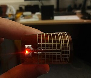 A printed rectenna lighting an LED from a Powercast 915 MHz transmitter, flexible meshed antenna bent with a red LED light