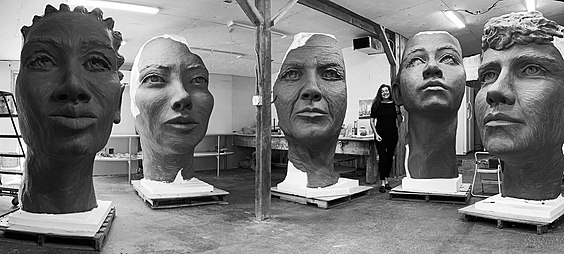 Black and white image of Amanda standing next to the 7' tall clay faces in her studio for The Girl Puzzle Monument in New York City honoring Nellie Bly.