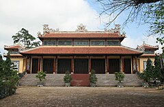 Un temple aux tuiles rouges, des piliers soutenant les deux niveaux.  Il y a des marches en béton menant au temple et des arbustes en pot devant eux.  Une cour en pierre est au premier plan, flanquée d'autres arbustes.