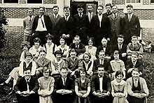 Photograph of four rows of men and women in formal-wear in their early-twenties sitting on a set of stairs with a brick building in the background