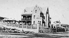 Die schwarz-weiß Fotografie im Querformat zeigt ein großes Haus mit Balkonen aus Holz umgeben von einem Zaun.