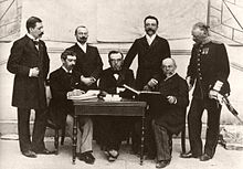 photographie noir et blanc : sept hommes trois assis à une table, quatre debout