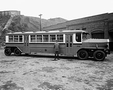 Pohled z dálky na dlouhý 8kolový městský autobus zaparkovaný před cihlovou budovou. Autobus je asi 1,5krát tak vysoký jako uniformovaný muž v popředí.