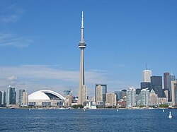 A spear-like tower is between a white-domed structure and small buildings on the left, and increasingly taller buildings to the right. In the foreground is a lake, with a few visible buoys, and the background is a deep blue sky with a few clouds near the horizon.