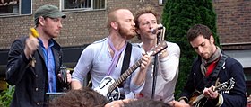 Una fotografía en color de cuatro miembros de Coldplay tocando con instrumentos frente a un edificio de ladrillo