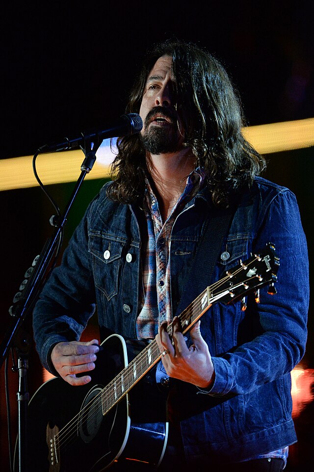 A man holding a guitar, wearing a blue shirt and a dark vest