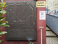 Gedenktafel mit Grundriss der Synagoge und Inschrift „An dieser Stelle stand die Ehrenfelder Synagoge, verbunden mit einer Religionsschule für Mädchen und Jungen, erbaut 1927 nach dem Entwurf des Architekten Robert Stern, zerstört am Tag nach der Reichspogromnacht am 9. November 1938“. Rote Hausfassade, neben der Gedenktafel direkt ein gelbes „Ausfahrt verboten“-Schild und ein Handzettel des körnerstrassenfests. Daneben ragt noch der Stiel eines Spatens ins Bild