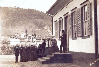 Fotografia mostrando um grupo de homens e mulheres ao pé da escada que leva a uma casa branca com persianas escuras