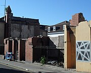 A red-brick building in the process of being demolished.  Stripped inner walls and a tall pinnacle are visible, and a metal fence and stubs of walls remain in the foreground.