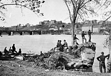 Black-and-white photo of several military men idling on a riverbank. Across the river are several large buildings.