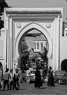 Schwarzweiss-Fotografie einer großen weißen Außentür mit orientalischen Formen.  Mehrere Personen und Fahrzeuge steigen durch die Tür ein und aus.