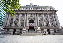 Uma imagem frontal baixa da Alfândega dos EUA de Alexander Hamilton.  Em frente ao prédio, há uma pequena escada com estátuas dos dois lados.  Existem várias janelas cobrindo a frente do edifício.