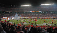 Далекие фигуры с оранжевыми флагами устремляются на затемненное футбольное поле. На переднем плане головы зрителей.