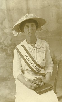 A photograph of Alice sitting on a chair wearing a sash that says Votes For Women, holding books