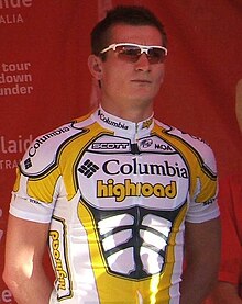 A man in his late twenties wearing a yellow and white cycling jersey with black trim and sunglasses on his head, standing calmly.