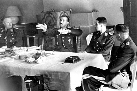 Black-and-white photograph of four men wearing uniforms sitting on wooden chairs around a table in a living room. An older man is sitting at the head of the table on the left. Two younger men are sitting along the table's side, with their backs towards a tiled fireplace on the room's far wall. A fourth man is sitting at the head of the table on the right, leaning back, his left leg folded over his right. The table is covered by a white table cloth. The right-hand side of the table is empty, except for a large dark ashtray, a spoon and an empty glass; the left and centre of the table is covered by an assortment of empty plates, coffee cups and other dishes; there is also a bowl of fruit. The second man from the left has his hands raised, palms facing the camera, and the fingers of both hands pointing to the left of the image. The heads of the three other men are turned towards him.