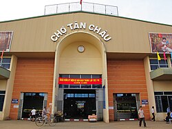 A large front entrance to a market, made from brick and sheet metal, in a modern style. The name of the market is above the front arch in large white letters, and the open interior can be faintly seen through the doors. People are walking in and out.