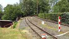 Dvě souběžně vedené úzkorozchodné železniční koleje. Vlevo od tratě lemované ostříhaným živým plotem z jehličnatých stromů. Mezi kolejemi a kolem nich železniční značky.