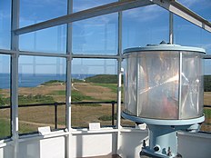 Foto prenita de la lanternĉambro de la lumturo.
En la malfono dekstraflanke estas la cirkla lenso ĉirkaŭanta la lampon.
La fono estas la vido atentanta super la lumturgrundoj kaj la klifo al la oceano.