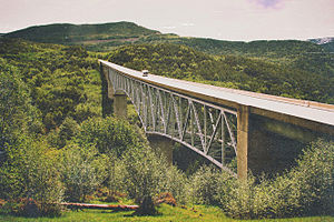 A steel bridge with a slight arch, carrying a two-lane road across a gorge that cuts between grasslands with small trees.