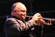 A 45-year-old man is shown in right profile, he is playing a trumpet with his right hand manipulating the valves and his left holding the instrument to his pursed lips. He has sparse thin head hair, his eyes are closed and he wears a brown pin-strip suit jacket. The front of the trumpet is out of shot.