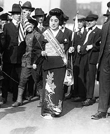 A black and white photograph taken in New York in 1917. A young Japanese woman wearing layered kimono stands in the centre, hold ing an American flag in her right hand and a Japanese flag in her left. She wears a traditional hairstyle and a diagonal sash that reads 