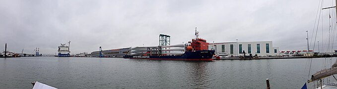 Livraison des pâles d'éoliennes en mer dans le port de Saint Nazaire. Au second plan, le navire Vestvind est à quai, chargé des pales d'éoliennes qui seront montées sur les turbines des éoliennes offshore. À l'arrière plan à gauche, le paquebot Celebrity Beyond en cours d'armement dans le bassin de Penhouët. À l'arrière plan à droite, l'usine de moteurs thermiques Man, et à l'extrême droite les turbines entreposées en attendant de repartir vers le champ en mer.