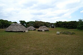 Photograph of the reconstructed Kaizuka Period village of the Nakabaru Site. It shows three huts with thatched roof in a clearing covered with grass, and another collapsed hut that permits to see the internal structure of the huts.