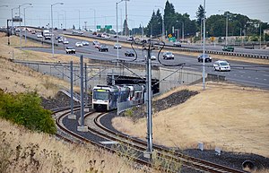 A light rail train with two cars enters a shallow, two-track tunnel under a freeway
