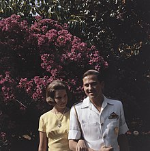 Photographie en couleur d'un couple se tenant par le bras, dont la femme porte une robe jaune et un collier de perles et l'homme porte un uniforme militaire blanc.