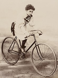 White woman posed on a bicycle, wearing a baggy white garment with a Union Jack on the chest