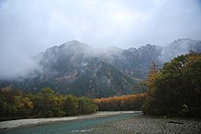 Fotografie color a unui munte înconjurat de nori, cu un râu la poalele sale.