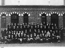 Photographie noir et blanc d'étudiants en quatre rangs avec leurs professeurs, devant un bâtiment en brique.