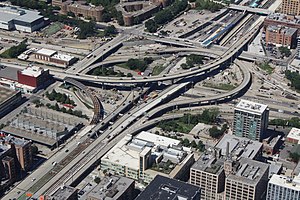 Aerial view of a major freeway interchange with several flyover ramps set within an urban neighborhood. Several ramps have exposed steel beams and other unfinished surfaces, showing signs of construction activity.