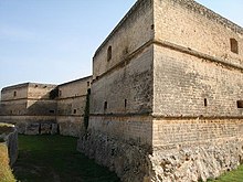 Una estructura de piedra de tres pisos con paredes lisas y una base de corte tosco.  Las paredes son angulares y tienen aberturas.