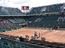 A red clay court in the middle of a stadium. No players are on the court and the bleachers are nearly empty