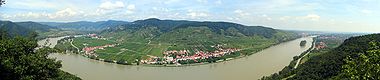 Ein riesiger Fluss schlängelt sich durch eine flache Landschaft. Am Ende der Flutebene erheben sich steile Berge über Häusern mit roten Dächern.