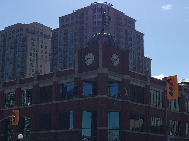 A three storey building of dark red brick stands in front of larger residential buildings. On the corner of the building at an intersection, a small tower one storey higher has a clock tower, and a copper statue on top of a copper dome