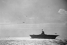 Photo en noir et blanc d'un porte-avions vu de bâbord sur lequel un avion apponte, pendant qu'un autre patrouille au-dessus.