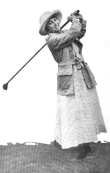 A smiling white woman swinging a golf club; she is wearing a brimmed hat, a cardigan, and a long white skirt