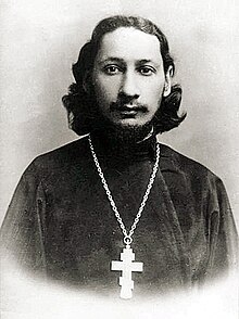 photographie d'un homme blanc avec les cheveux longs en tenue religieuse, une grosse croix autour du coup.