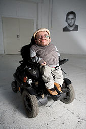 Zu sehen ist Raul Krauthausen in seinem Rollstuhl sitzend. Der Rollstuhl ist ein elektronischer und in dunklen Farben gehalten. Raul K.trägt beige Kleidung und eine helle Kappe.gtn