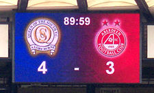 A digital scoreboard displaying the elapsed time of a football match and the club crests of the competitors, with their respective scores below.